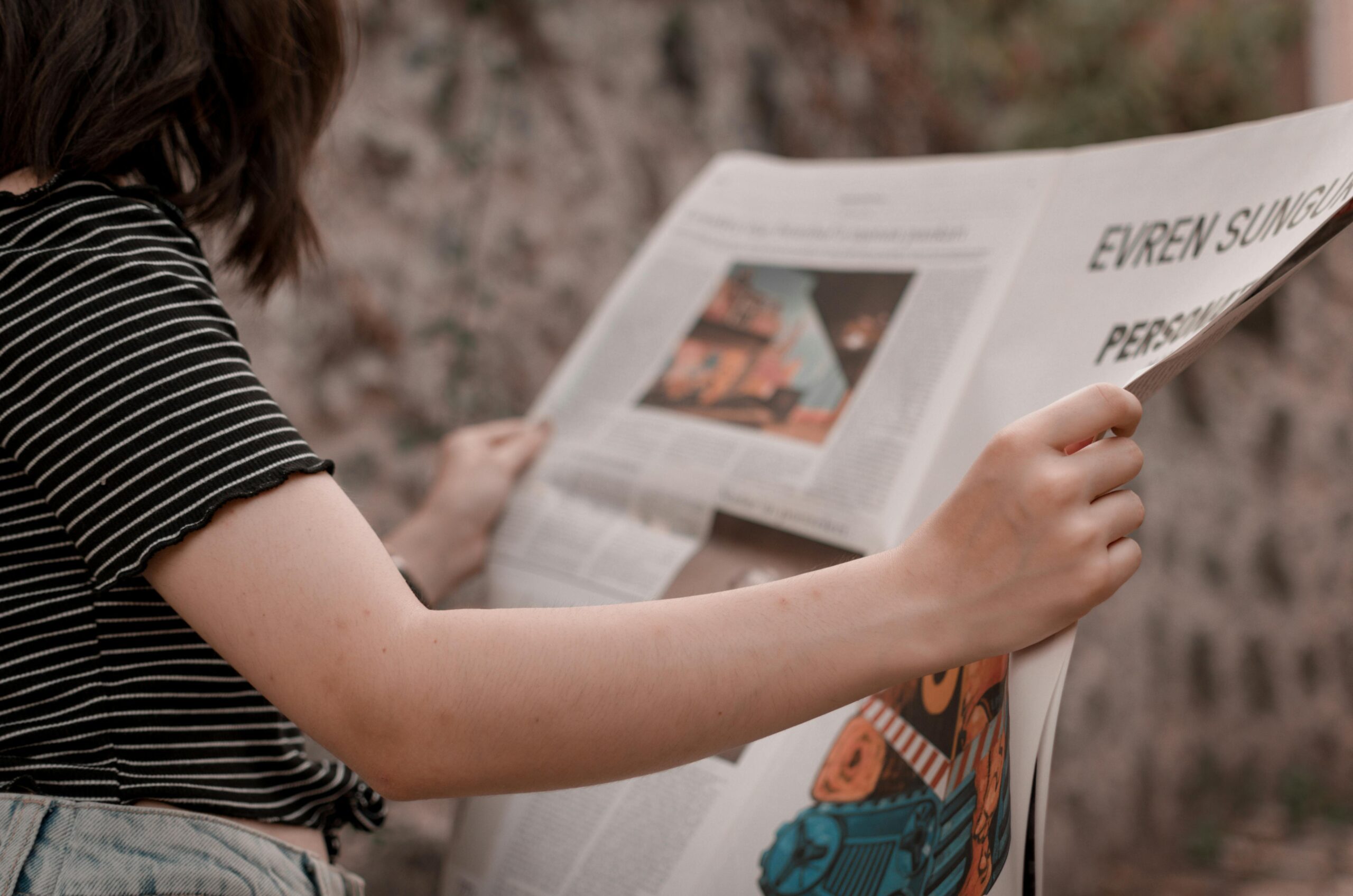 A woman holding a newspaper while sitting outside, enjoying a leisurely read.